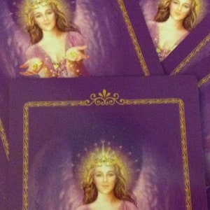 May include: Purple tarot cards featuring an angelic figure with a golden crown and wings. The angel wears a pink dress and holds glowing orbs in her hands. The cards have a gold border and are stacked.