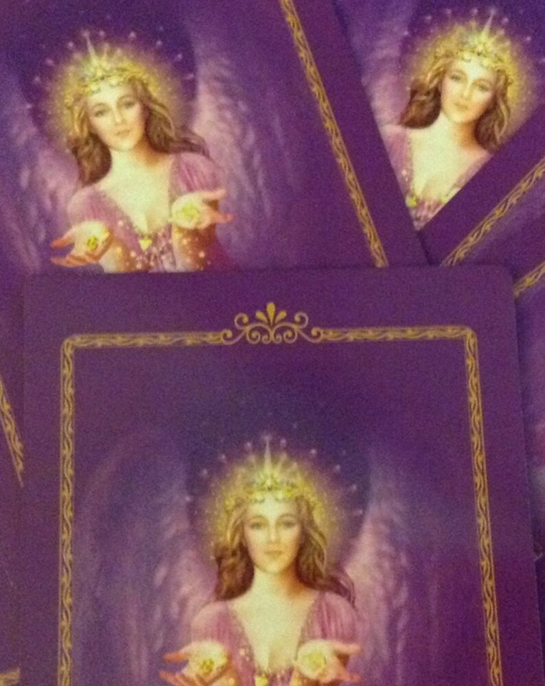 May include: Purple tarot cards featuring an angelic figure with a golden crown and wings. The angel wears a pink dress and holds glowing orbs in her hands. The cards have a gold border and are stacked.
