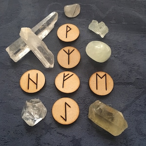 May include: An assortment of crystals and wooden rune stones arranged on a dark blue textured surface. Clear quartz crystals, various shapes of gemstones, and wooden rune stones with black symbols are visible.