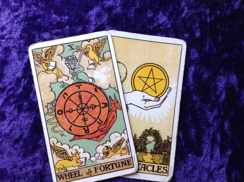 May include: Two tarot cards are displayed, featuring colorful illustrations. The "Wheel of Fortune" card shows a wheel with symbols and text, while the "Pentacles" card depicts a hand holding a golden pentacle. The cards are set against a textured purple background.