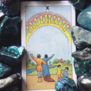 Op de afbeelding: Een tarotkaart met een familiescène onder een regenboog van tien bekers. De kaart toont figuren in kleurrijke kleding, met een landelijke achtergrond. De kaart is omgeven door blauwe en groene kristallen.