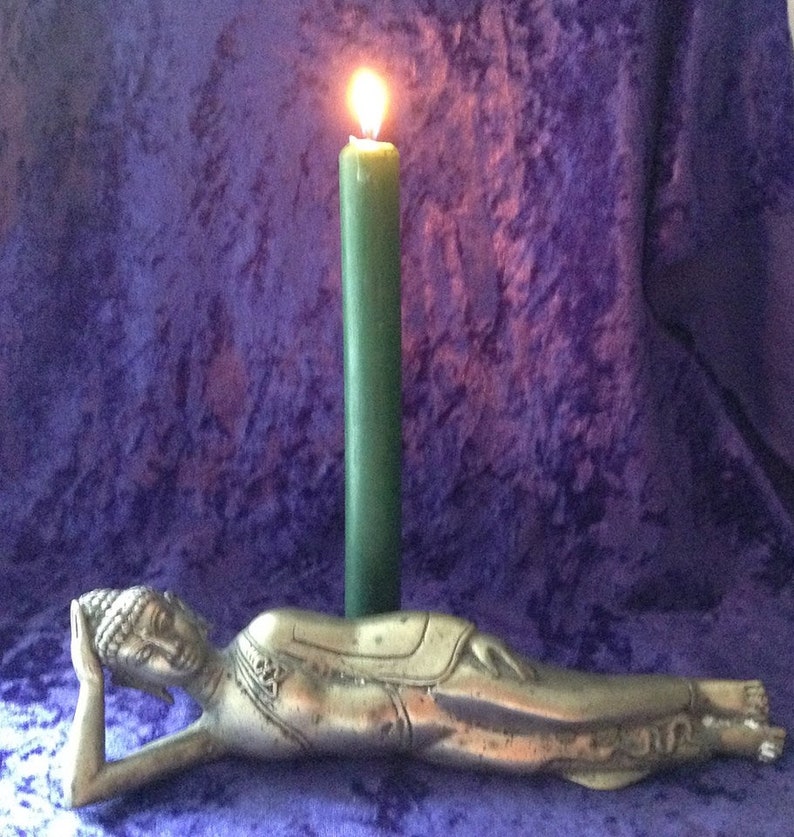K&ouml;nnte beinhalten: Eine silberne liegende Buddha-Statue mit einer brennenden gr&uuml;nen Kerze davor. Die Statue liegt auf einem violetten Samtstoff.