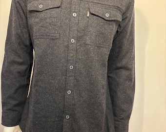May include: A dark gray button-down shirt with two chest pockets and a small tag on the right pocket. The long-sleeved shirt has a collared neckline and is displayed on a mannequin.