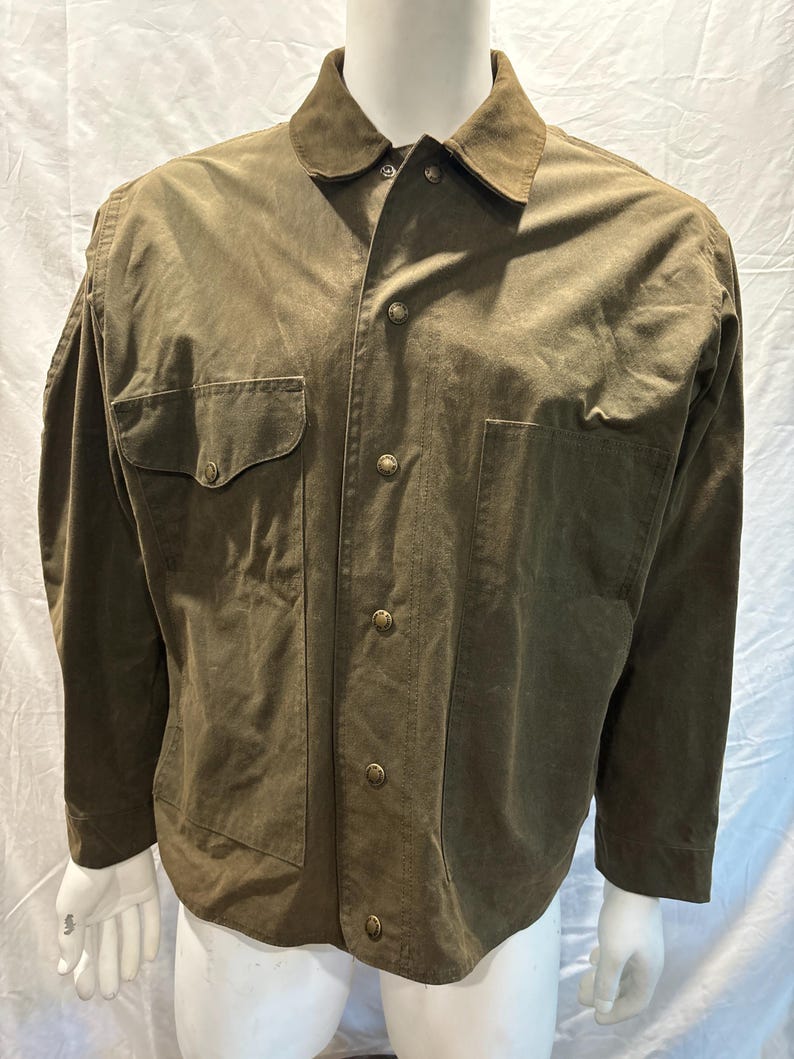 Puede incluir: Una camisa de manga larga de color verde oliva con cuello de ante y varios bolsillos. La camisa tiene cierre de botones en la parte delantera y botones de lat&oacute;n.