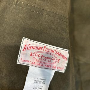 Puede incluir: Una etiqueta blanca con texto negro que dice "A Genuine Filson Garment" y "C-C-Filson Co. Seattle" en un c&iacute;rculo con flechas que apuntan hacia afuera. La etiqueta tambi&eacute;n dice "Might as well have the best". Debajo de la etiqueta hay una etiqueta blanca m&aacute;s peque&ntilde;a con texto negro que dice "C.C. Filson Seattle, WA Made in USA Style 420 9711 7306 Size XL 100% Cotton Verse".