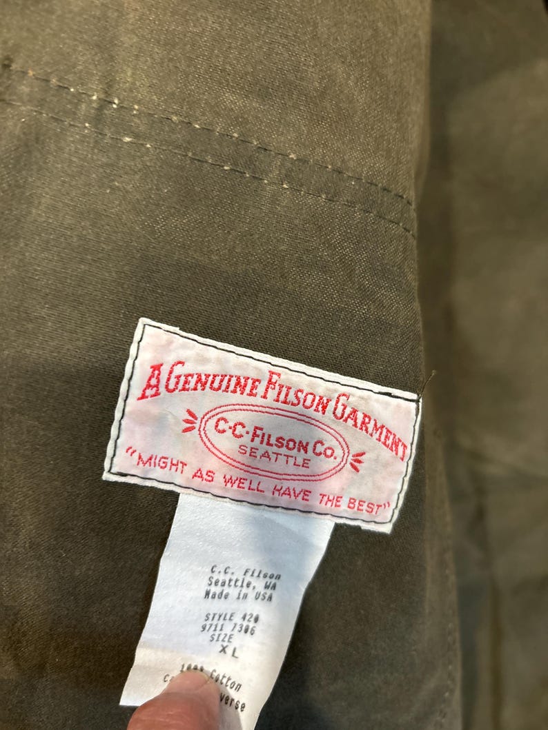 Puede incluir: Una etiqueta blanca con texto negro que dice "A Genuine Filson Garment" y "C-C-Filson Co. Seattle" en un c&iacute;rculo con flechas que apuntan hacia afuera. La etiqueta tambi&eacute;n dice "Might as well have the best". Debajo de la etiqueta hay una etiqueta blanca m&aacute;s peque&ntilde;a con texto negro que dice "C.C. Filson Seattle, WA Made in USA Style 420 9711 7306 Size XL 100% Cotton Verse".