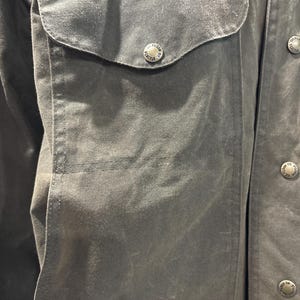 Puede incluir: Primer plano de una chaqueta de algod&oacute;n verde oscuro con un gran bolsillo de solapa en el lado izquierdo. El bolsillo tiene un bot&oacute;n con las palabras "Filson Seattle" grabadas.