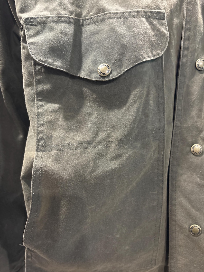 Puede incluir: Primer plano de una chaqueta de algod&oacute;n verde oscuro con un gran bolsillo de solapa en el lado izquierdo. El bolsillo tiene un bot&oacute;n con las palabras "Filson Seattle" grabadas.