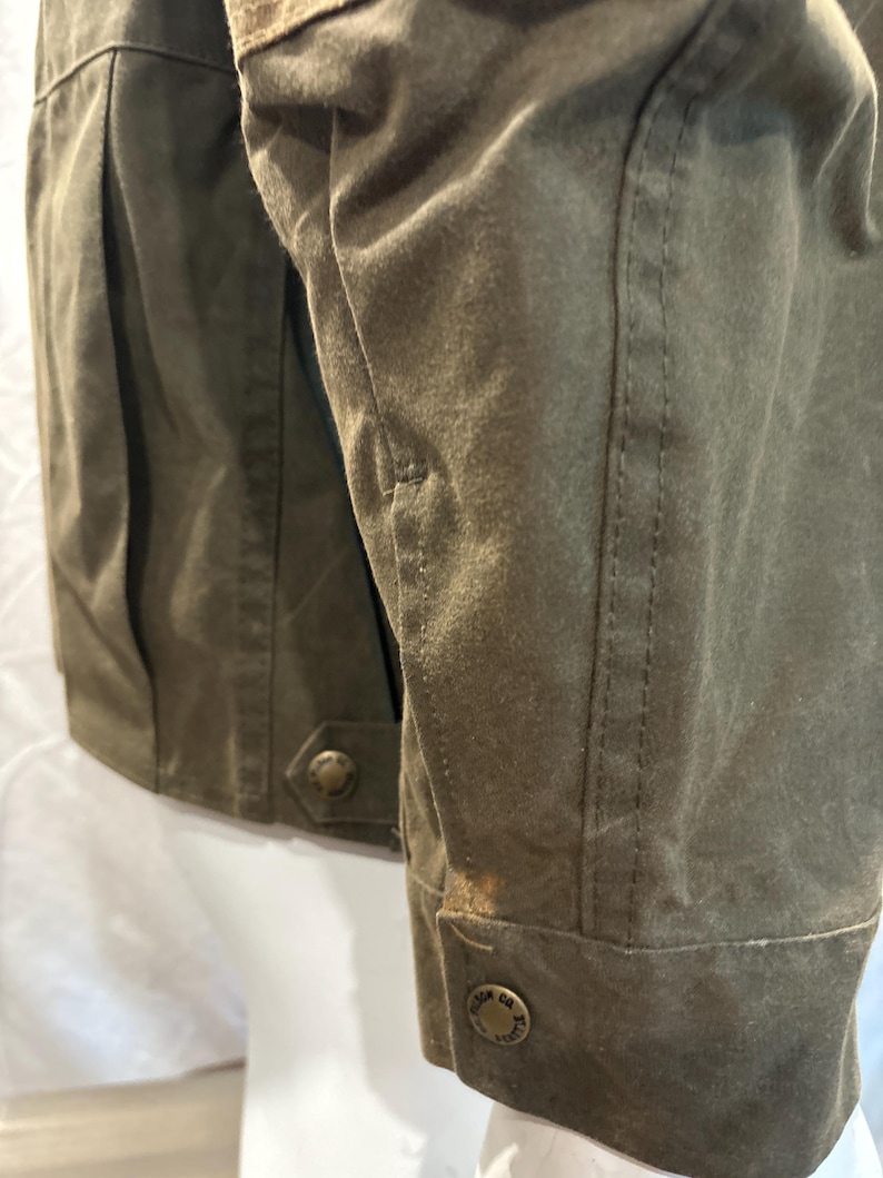 Puede incluir: Una chaqueta de lona verde con un parche de cuero marr&oacute;n en la manga. La chaqueta tiene cierre de botones y un bolsillo con bot&oacute;n en el lado izquierdo. La chaqueta tiene una etiqueta que dice "Field Co. Sports" en la parte inferior de la manga.