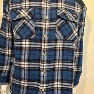 May include: A long-sleeve button-down shirt featuring a blue, black, and white plaid pattern. The shirt includes solid black shoulder panels, a standard collar, and two chest pockets with buttoned flaps. The front is secured with light-colored buttons.