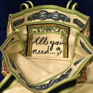 May include: A canvas tote bag with an open top and two handles. The interior features a cream-coloured lining with a green and black striped trim. A central pocket displays the text All you need... with a small flower illustration and a bow motif.