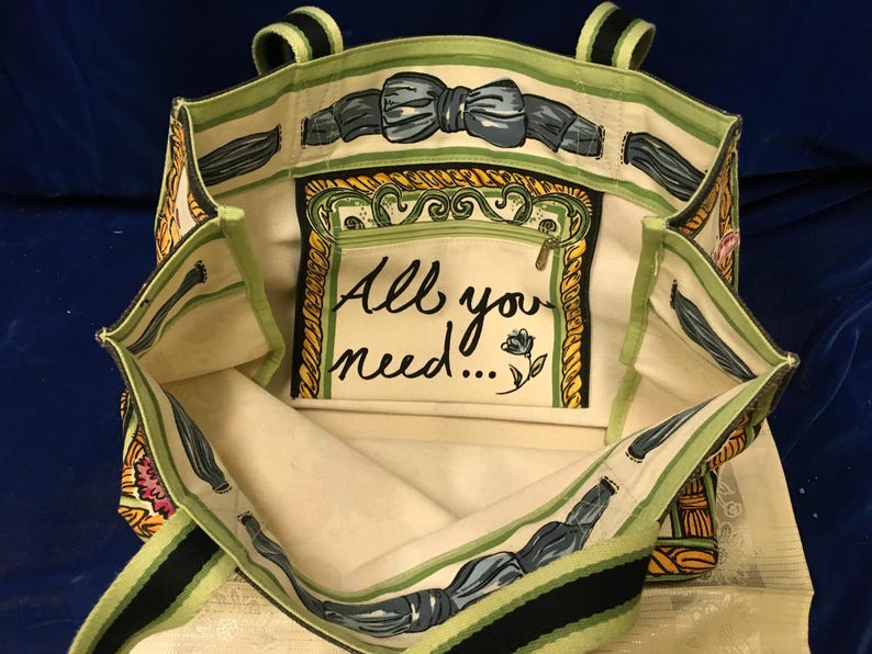 May include: A canvas tote bag with an open top and two handles. The interior features a cream-coloured lining with a green and black striped trim. A central pocket displays the text All you need... with a small flower illustration and a bow motif.
