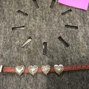 Puede incluir: Una pulsera de cuero marr&oacute;n con cuatro dijes plateados en forma de coraz&oacute;n. Los dijes tienen dise&ntilde;os intrincados y est&aacute;n espaciados uniformemente a lo largo de la pulsera. La pulsera tiene una hebilla plateada y una punta blanca.