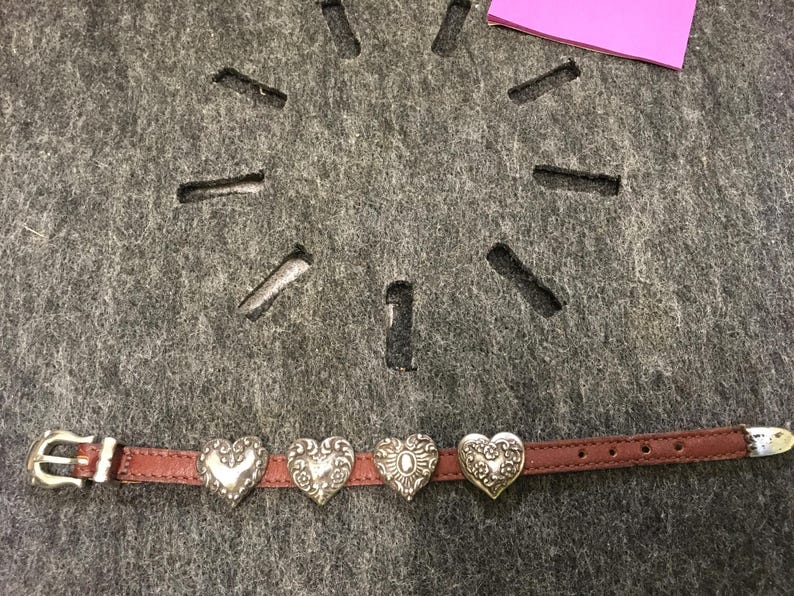 Puede incluir: Una pulsera de cuero marr&oacute;n con cuatro dijes plateados en forma de coraz&oacute;n. Los dijes tienen dise&ntilde;os intrincados y est&aacute;n espaciados uniformemente a lo largo de la pulsera. La pulsera tiene una hebilla plateada y una punta blanca.
