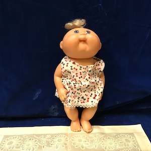 May include: A doll with a round face, blue eyes, and a small tuft of blonde hair tied at the top. The doll wears a sleeveless white dress featuring a repeating pattern of small red and green bows, finished with a delicate white lace trim at the hem.