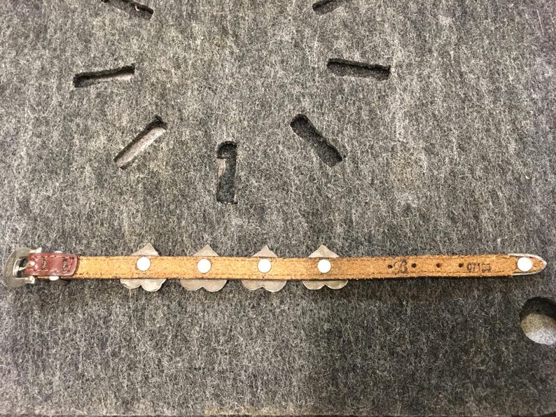 Puede incluir: Un cintur&oacute;n estrecho de cuero marr&oacute;n claro con detalles decorativos plateados. El cintur&oacute;n presenta una peque&ntilde;a hebilla plateada y una serie de piezas met&aacute;licas en forma de coraz&oacute;n con tachuelas circulares blancas. El cintur&oacute;n est&aacute; sobre una superficie gris.