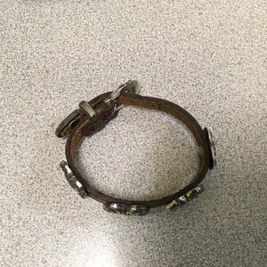 Puede incluir: Una pulsera de cuero marr&oacute;n con detalles met&aacute;licos plateados. La pulsera tiene un cierre de hebilla y piezas met&aacute;licas decorativas. La pulsera es circular y parece sin usar.