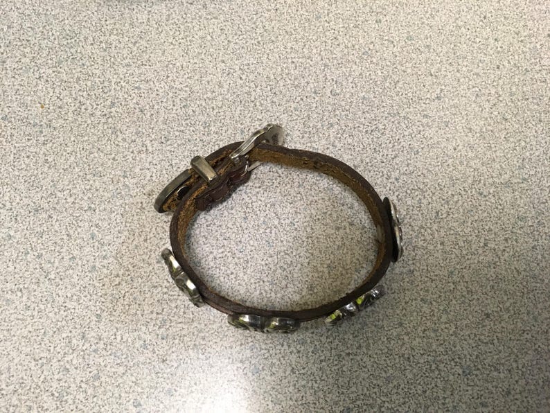 Puede incluir: Una pulsera de cuero marr&oacute;n con detalles met&aacute;licos plateados. La pulsera tiene un cierre de hebilla y piezas met&aacute;licas decorativas. La pulsera es circular y parece sin usar.