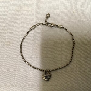 May include: Silver bracelet featuring a heart-shaped pendant. The pendant has a heart-shaped cutout. The bracelet is displayed on a white, textured surface. The chain is a box chain style.