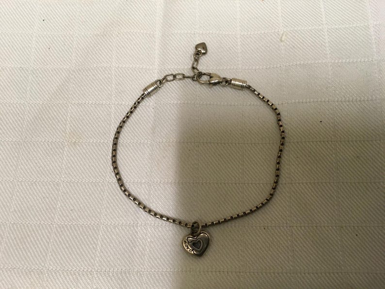 May include: Silver bracelet featuring a heart-shaped pendant. The pendant has a heart-shaped cutout. The bracelet is displayed on a white, textured surface. The chain is a box chain style.