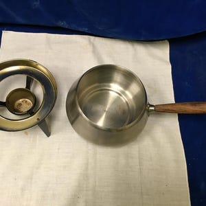 Può includere: Un pentolino per fondue in acciaio inossidabile con un manico in legno e un supporto separato in metallo dorato con tre gambe.