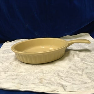 以下が含まれることがあります： 白いテーブルクロスの上に置かれた、ベージュのセラミック製のベーキングディッシュ。ハンドルが1つ付いており、縁はフリル状になっている。