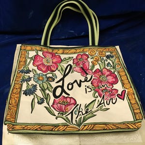 May include: A cream-coloured tote bag featuring a floral print with pink, blue, and yellow flowers, a butterfly, and a small red heart. The text "Love is in the Air" is printed in black script. The bag has a yellow and black woven-style border.