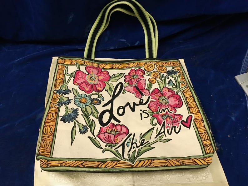 May include: A cream-coloured tote bag featuring a floral print with pink, blue, and yellow flowers, a butterfly, and a small red heart. The text "Love is in the Air" is printed in black script. The bag has a yellow and black woven-style border.