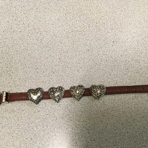 Puede incluir: Un cintur&oacute;n de cuero marr&oacute;n con cuatro adornos plateados en forma de coraz&oacute;n. El cintur&oacute;n tiene una hebilla y una punta plateadas. Los corazones tienen dise&ntilde;os detallados y est&aacute;n espaciados uniformemente a lo largo del cintur&oacute;n. Mide unos 100 cm.