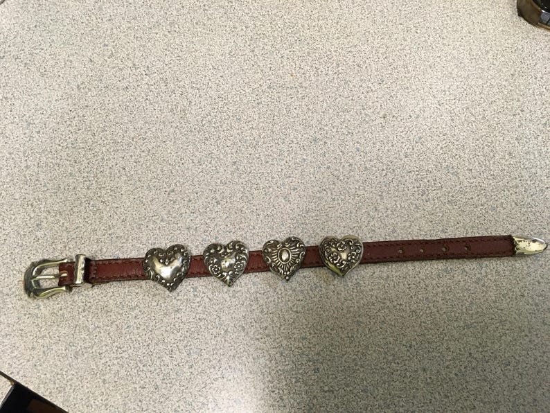 Puede incluir: Un cintur&oacute;n de cuero marr&oacute;n con cuatro adornos plateados en forma de coraz&oacute;n. El cintur&oacute;n tiene una hebilla y una punta plateadas. Los corazones tienen dise&ntilde;os detallados y est&aacute;n espaciados uniformemente a lo largo del cintur&oacute;n. Mide unos 100 cm.