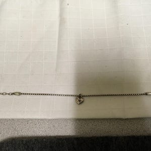 May include: A silver bracelet featuring a fine chain and a heart-shaped charm. The charm has a textured appearance. The bracelet is displayed on a white, textured background.