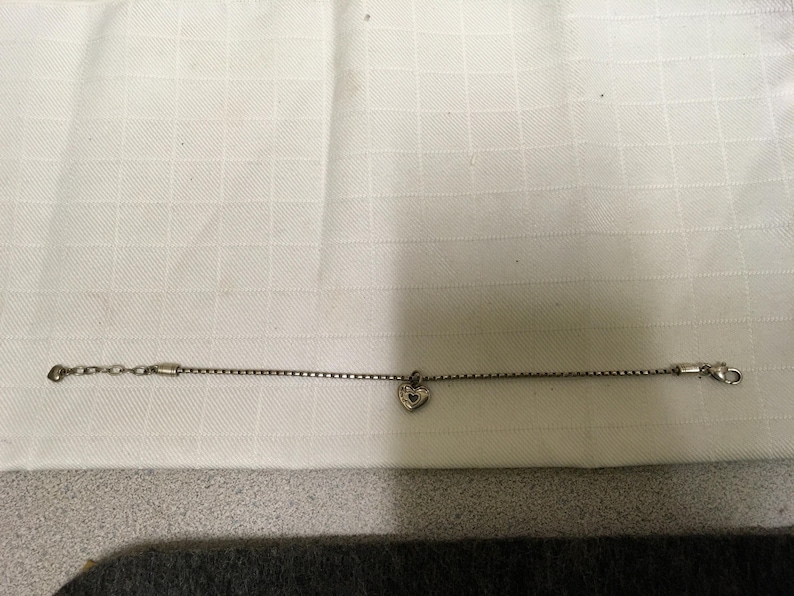 May include: A silver bracelet featuring a fine chain and a heart-shaped charm. The charm has a textured appearance. The bracelet is displayed on a white, textured background.