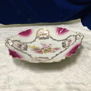 May include: An oval antique serving dish with a scalloped edge. The dish has a white base with a floral design in the centre, accented by pink and gold trim. Decorative elements include a chain-like border.