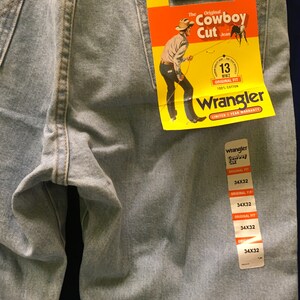 May include: A pair of light blue denim jeans with a yellow and red Wrangler tag attached. The tag says "The Original Cowboy Cut Jean" and features a cartoon cowboy. The jeans are size 34x32.