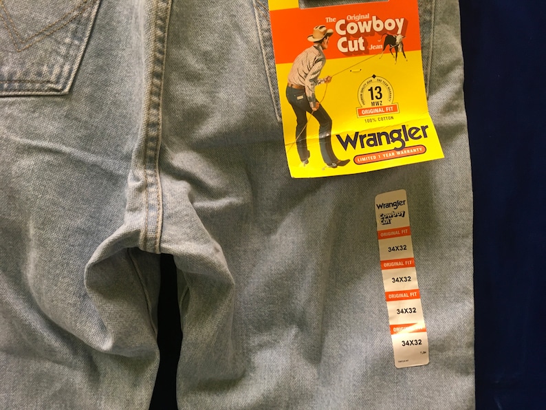 May include: A pair of light blue denim jeans with a yellow and red Wrangler tag attached. The tag says "The Original Cowboy Cut Jean" and features a cartoon cowboy. The jeans are size 34x32.