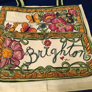 May include: A white canvas tote bag featuring a colourful illustration of pink flowers, orange and yellow butterflies, and a green woven-style border. The word Brighton is written in large black cursive script across the front pocket area.