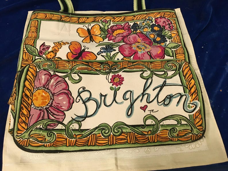 May include: A white canvas tote bag featuring a colourful illustration of pink flowers, orange and yellow butterflies, and a green woven-style border. The word Brighton is written in large black cursive script across the front pocket area.