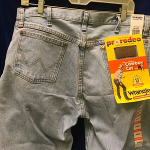May include: Light blue denim jeans with a brown leather Wrangler label and a yellow tag that reads "The Original Cowboy Cut" with a Wrangler logo.