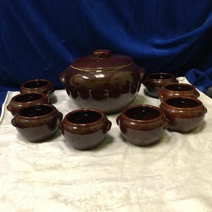 May include: A brown ceramic soup tureen with a lid, surrounded by eight matching soup bowls. The tureen and bowls have a glossy finish and small handles. The set is arranged on a white tablecloth.