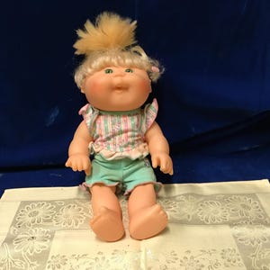 May include: A doll with blonde hair styled in a high tuft, green eyes, and rosy cheeks. The doll wears a sleeveless top with a light green, pink, and white striped pattern featuring small floral accents, paired with solid light green shorts.