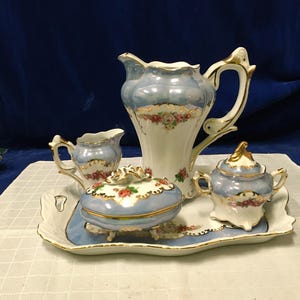 Puede incluir: Un juego de té de porcelana vintage en azul claro y blanco, adornado con motivos florales y detalles dorados. El juego incluye una tetera, una jarra de crema, un azucarero y una bandeja de servicio. Las piezas están sobre un mantel blanco.