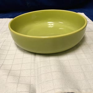 Peut inclure: Un bol rond en céramique vert clair avec un bord légèrement incurvé. Le bol est posé sur un tissu blanc à carreaux. Le bol est vide et semble neuf.