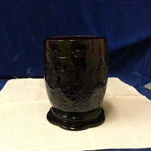 Puede incluir: Un vaso de vidrio burdeos oscuro con un patrón floral. El vaso tiene una base ancha y una parte superior ligeramente acampanada. El diseño floral está en relieve en la superficie del vidrio. El vaso está sobre una superficie blanca.