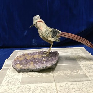 May include: A carved bird figurine with a crest, a long reddish-brown tail, and grey and white plumage, standing on a base of purple amethyst crystals. The bird has a sharp beak and a red streak above its eye, mounted on a natural mineral cluster.