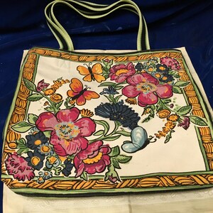 May include: A rectangular tote bag featuring a white base with a vibrant floral and butterfly print. The design includes large pink and dark blue flowers, green leaves, and orange butterflies, all framed by a yellow and green woven-style border.