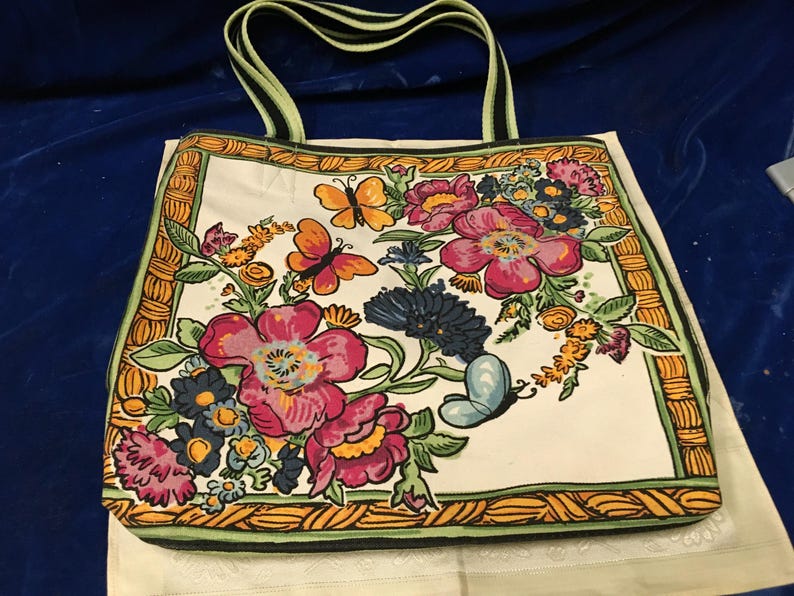 May include: A rectangular tote bag featuring a white base with a vibrant floral and butterfly print. The design includes large pink and dark blue flowers, green leaves, and orange butterflies, all framed by a yellow and green woven-style border.
