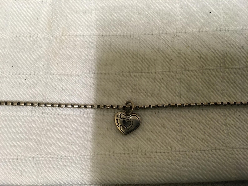 May include: Silver necklace featuring a heart pendant. The pendant has a heart outline with a smaller heart inside. The word "LOVE" is inscribed on the pendant. The chain is a box chain style.