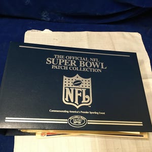 以下が含まれることがあります： 「THE OFFICIAL NFL SUPER BOWL PATCH COLLECTION」と銀色の文字とNFLのロゴが入った濃紺のバインダー。アメリカのプレミアスポーツイベントを記念しています。バインダーは白い布の上に置かれています。