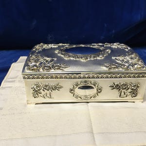 Puede incluir: Una caja rectangular para pañuelos de color plateado con motivos de rosas en relieve en la parte superior y frontal. El diseño incluye una abertura ovalada central en la parte superior y un elemento decorativo ovalado más pequeño en el frente, bordeado por un patrón de corazones.