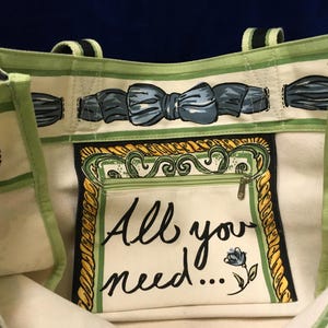 May include: A cream-coloured tote bag with light green trim and a blue bow pattern along the top edge. The interior features a central zippered pocket with a yellow rope-style border and the black printed text "All you need..." with a small blue flower.